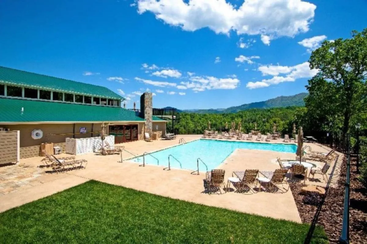 A spacious outdoor community pool area with inviting deck chairs, green grassy surroundings, and a panoramic mountain view. The pool is perfect for leisurely swims, sunbathing, or community gatherings.