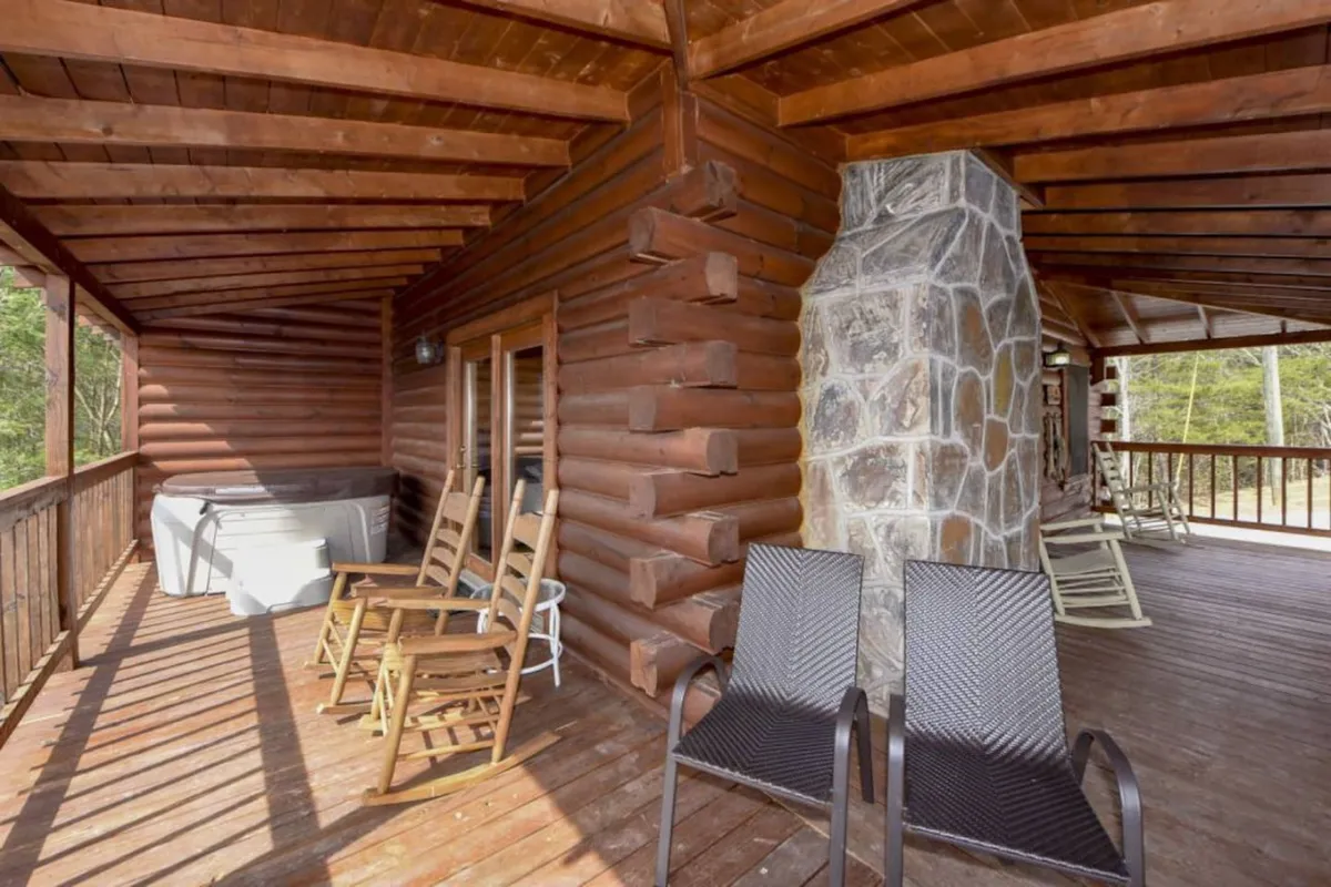 An inviting exterior porch featuring comfortable rocking chairs, a bubbling hot tub, and a charming stone fireplace. Surrounded by natural beauty, the porch offers a peaceful setting to unwind and connect with loved ones, with views of the mountains in the distance.