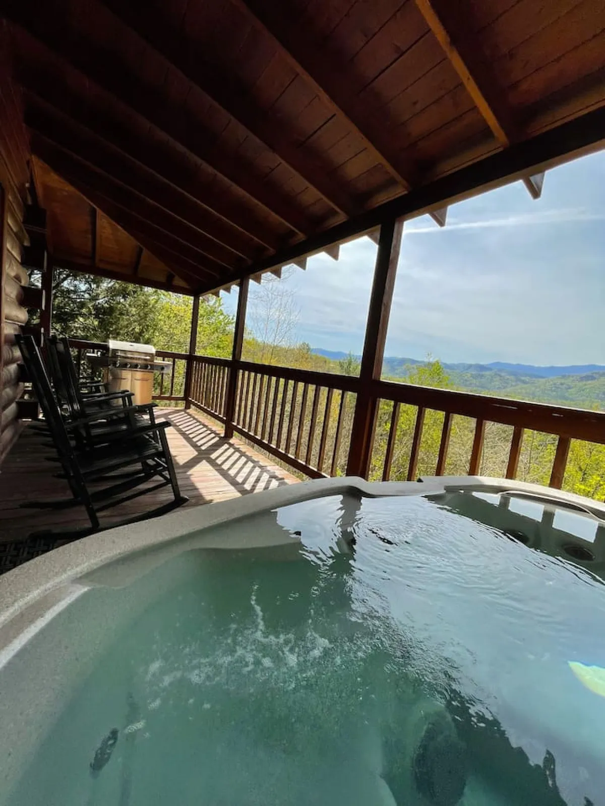 A cozy exterior deck with a soothing hot tub and wooden rocking chairs, all facing a spectacular mountain view. The scene promises relaxation after a day of hiking, with the option to soak in the hot tub while gazing at the sunrise or sunset over the peaks.