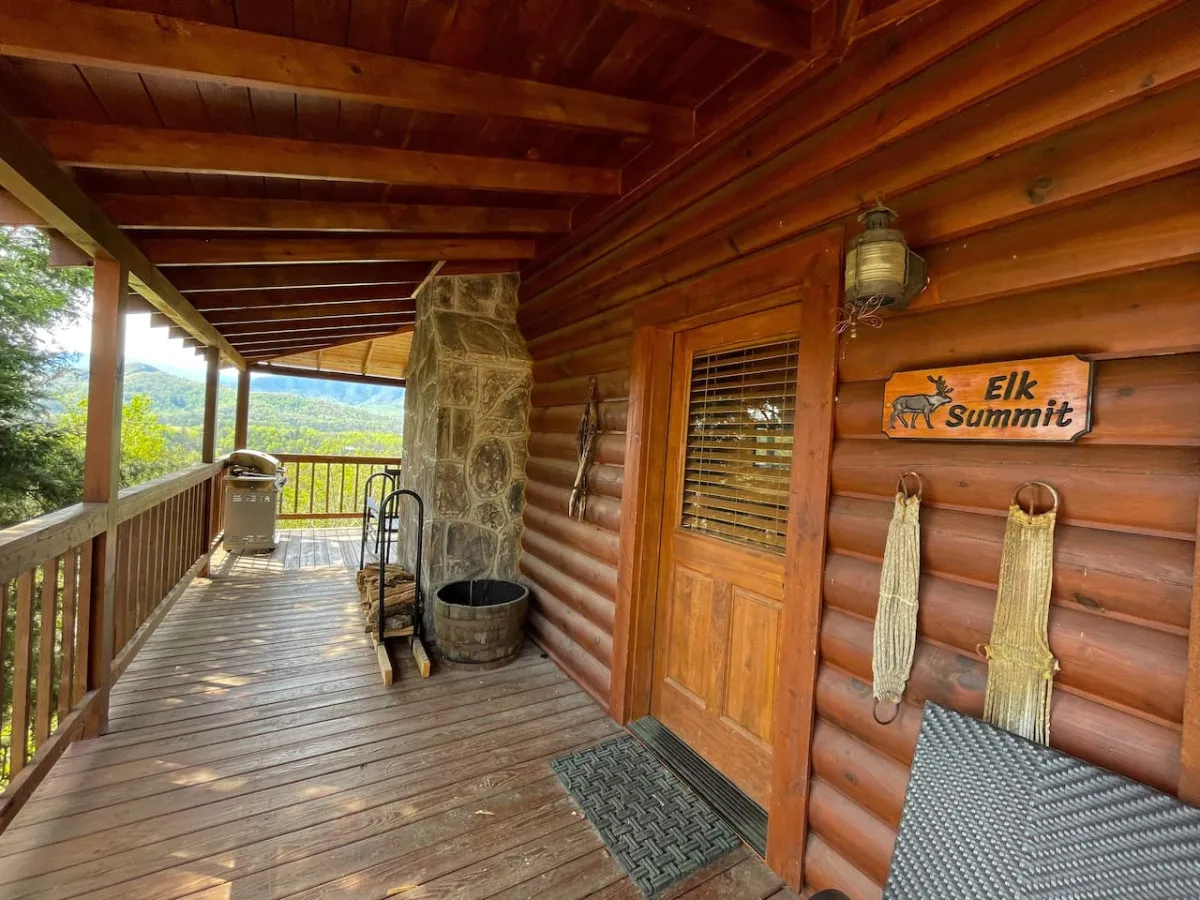 An inviting cabin entrance with a rustic wooden door, a stone chimney, and a scenic mountain view beyond the porch. The setting hints at tranquil mornings with coffee or evenings sharing stories, all with the mountains as a picturesque backdrop.