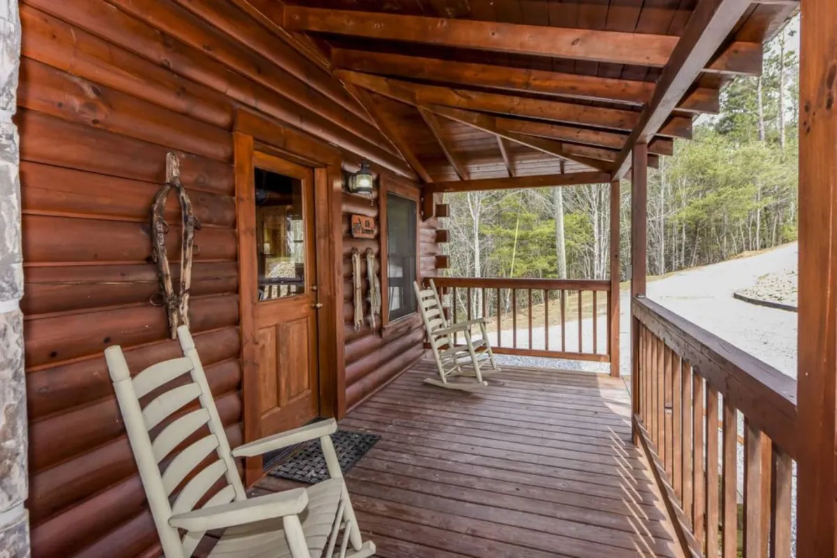 A cozy wooden porch with rocking chairs and a front door, bordered by a rustic railing. This welcoming space suggests a transition zone between indoors and outdoors—perfect for relaxing after a walk. Sunlight illuminates the area, and the calm atmosphere invites guests and their pets to unwind together.