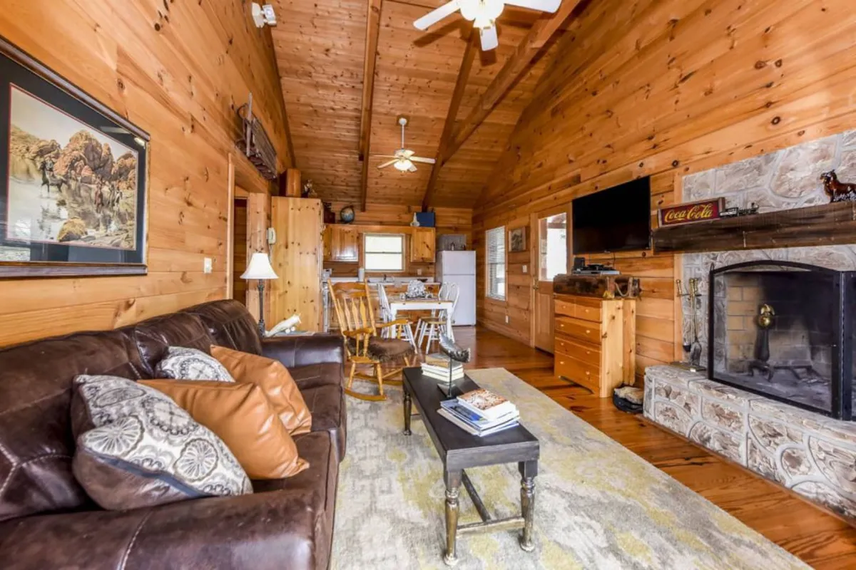 A warm and welcoming living room featuring a brown leather sofa, a rustic stone fireplace, a dining table with wooden chairs, and a TV mounted on the wall. Large windows allow natural light to flood the space, accentuating the inviting atmosphere and offering a glimpse of the outdoors.