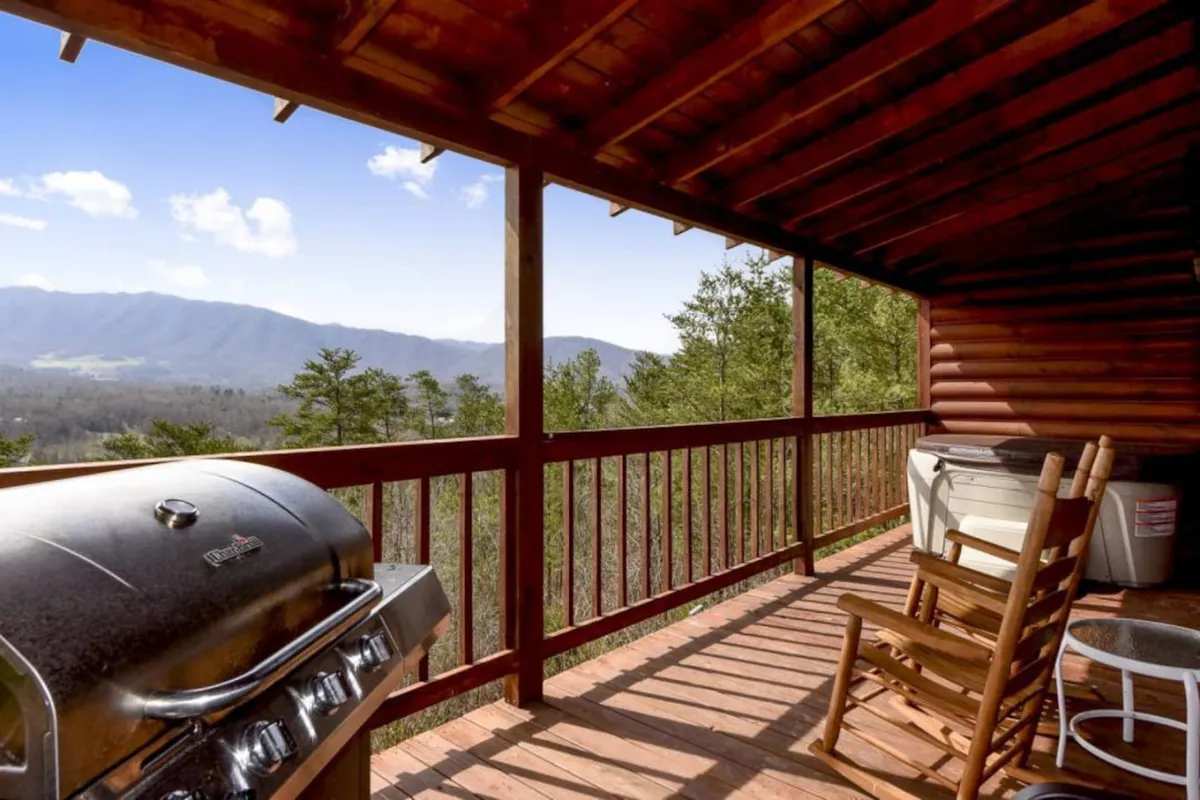 A spacious wooden deck with a grill and rocking chairs overlooks a stunning mountain landscape. The golden hues of sunset illuminate the sky, creating a romantic and peaceful setting. The porch’s wooden railing frames the panoramic views, hinting at the privacy and tranquility of the retreat.