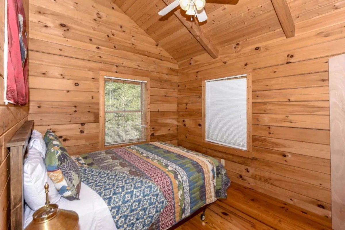 A cozy bedroom featuring a queen bed adorned with a colorful quilt, surrounded by warm wooden walls and natural light pouring in from windows that overlook the surrounding woodland.