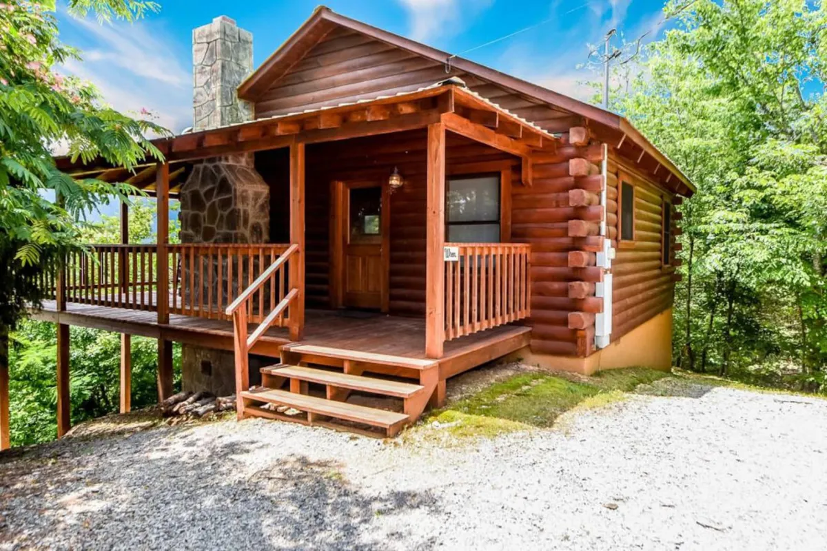 The image showcases a charming log cabin nestled among lush greenery, with a welcoming wooden porch, a rustic stone chimney, and panoramic views of the Smoky Mountains. The scene exudes warmth and tranquility, inviting guests to relax on the porch, take in stunning sunsets, and share memorable moments with family or friends.
