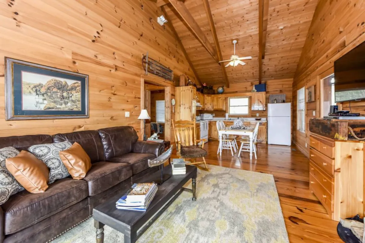A warm and inviting den area with a leather sofa, wooden kitchen cabinets, a dining area, and artwork, offering plenty of space to relax, entertain, or accommodate extra guests.