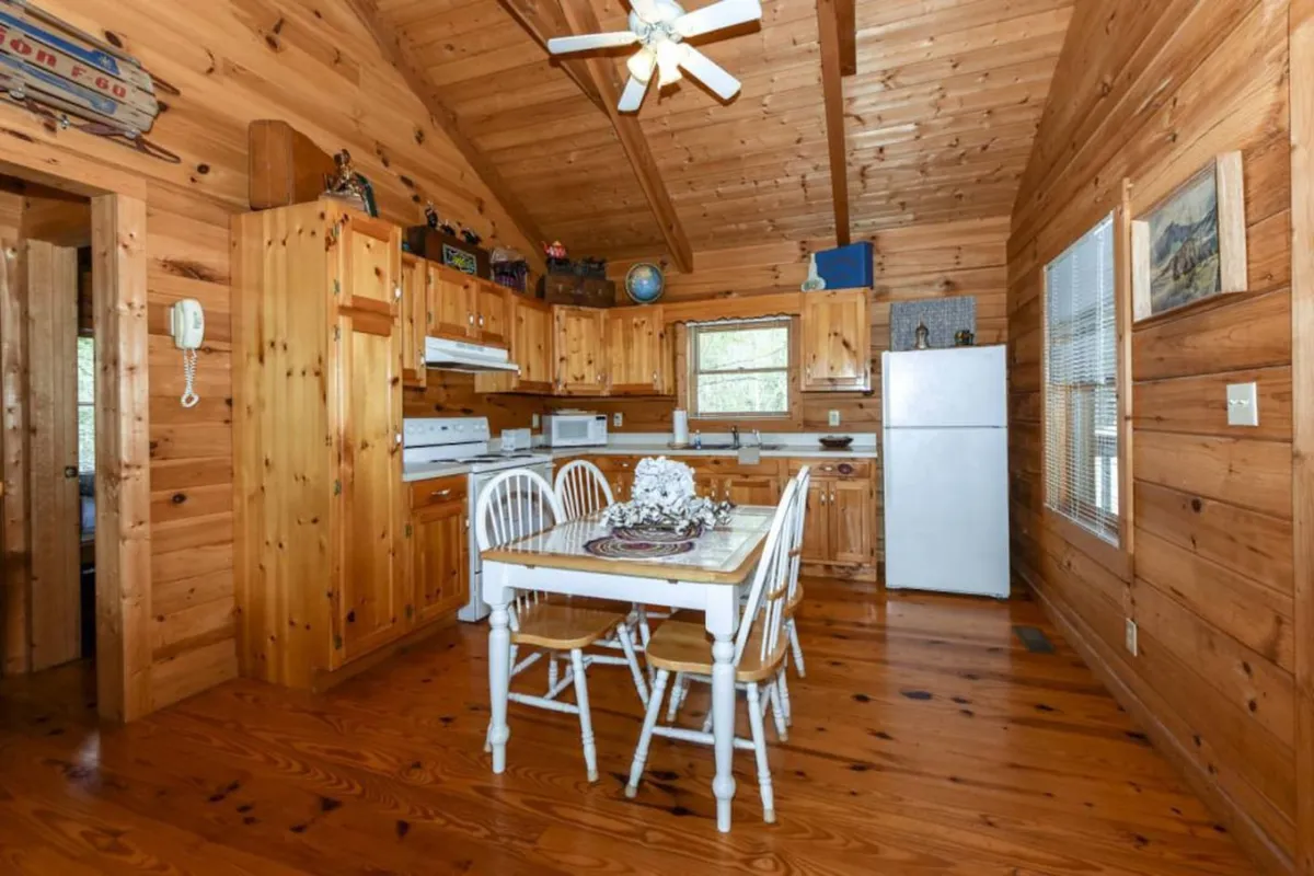 A warm, inviting kitchen and dining area featuring natural wood walls, wooden cabinetry, a central dining table, and full-sized kitchen appliances. The scene is set for family meals, group gatherings, or preparing food, with plenty of natural light and a cozy mountain cabin atmosphere.
