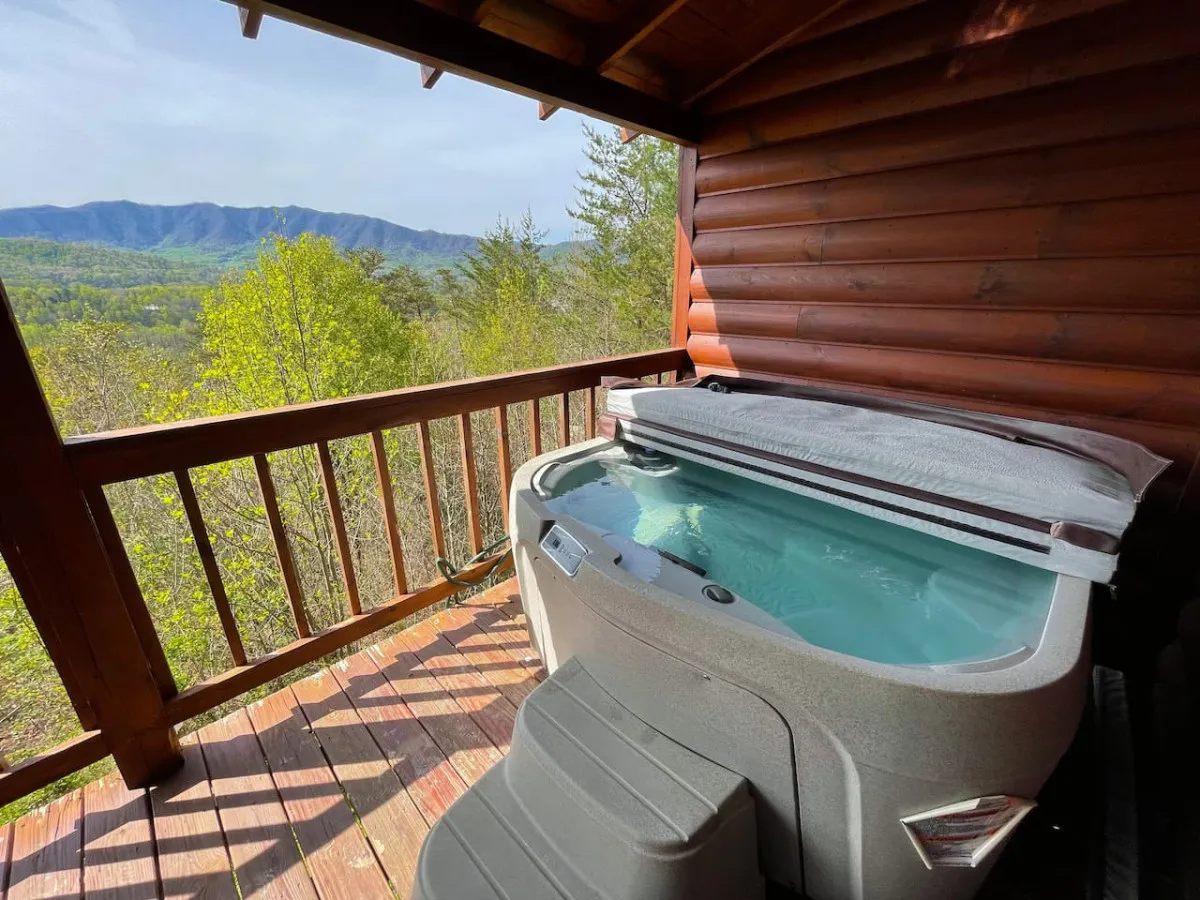 A private wooden deck featuring a bubbling hot tub with unobstructed views of the surrounding mountains and lush greenery. The tranquil setting is accentuated by the natural beauty, perfect for relaxation and unwinding outdoors.