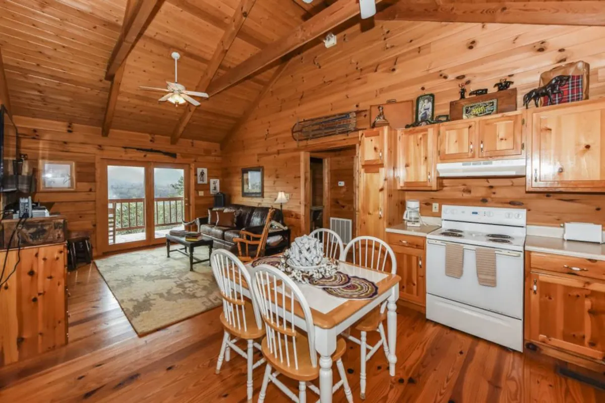 An interior view with wooden ceiling beams, a spacious kitchen island, comfortable seating area, and large windows framing scenic mountain vistas. The natural light and open layout foster a sense of connection between the indoor living space and the outdoors.