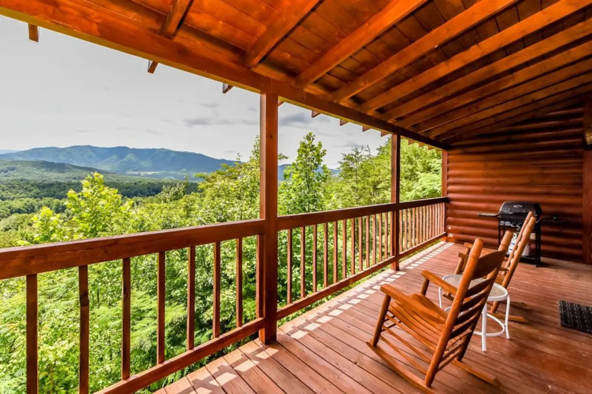 An exterior deck scene with wooden railings, rocking chairs, a grill, and an expansive mountain view. The outdoor grill is ready for barbecuing, and the space is ideal for alfresco dining while enjoying scenic vistas.